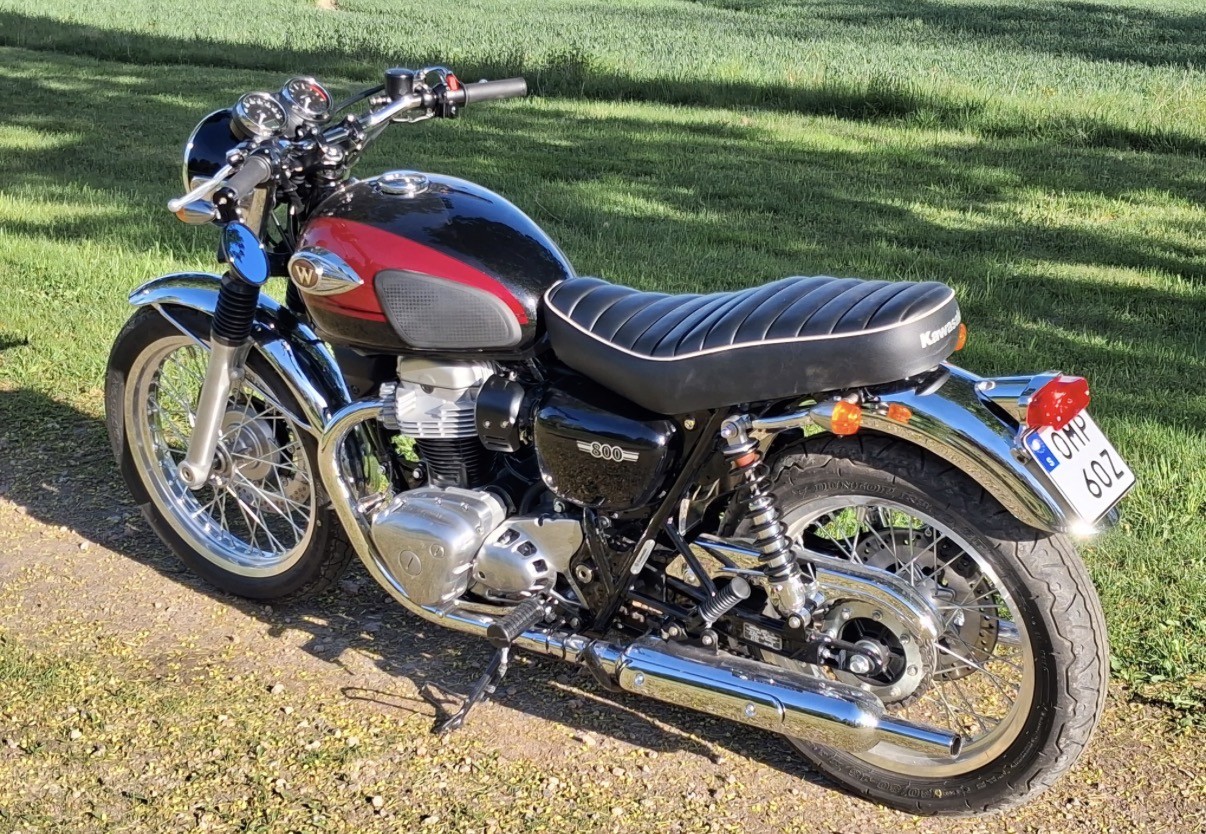 Svart och röd motorcykel av märket Kawasaki W800 med kromdetaljer parkerad på grusväg, gräsmatta i bakgrunden.